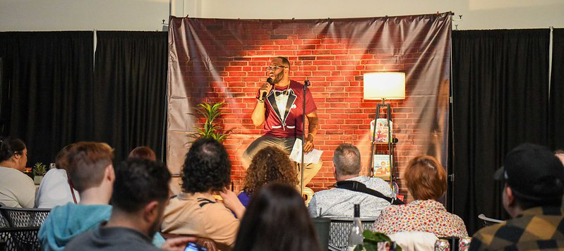 A comedian on stage talking to audience at Laughs for Literacy 2023 at the Chatsworth office.