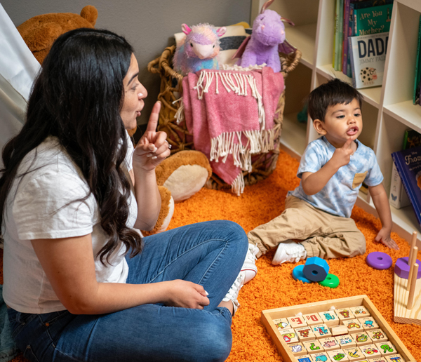 A child care provider and a child playing with toys on a orange rug.