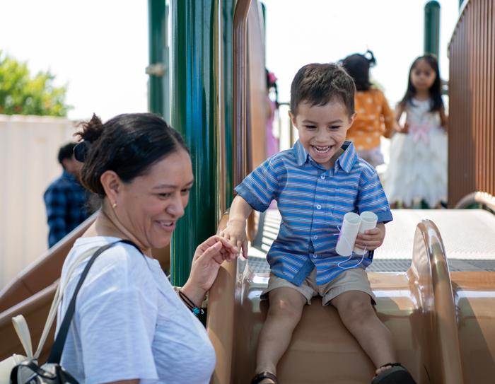 A boy smiling while riding down a slide and holding his mother's hand.