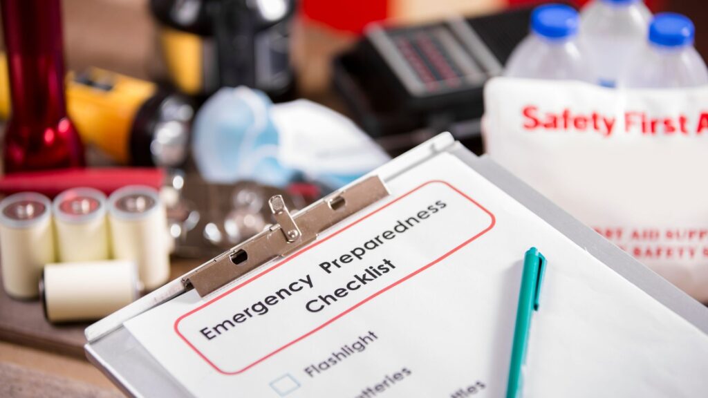 An image of a clipboard with the headline “Emergency Preparedness Checklist” and a pen on top of the clipboard and various safety objects in the background like water bottles, flashlights, and batteries.