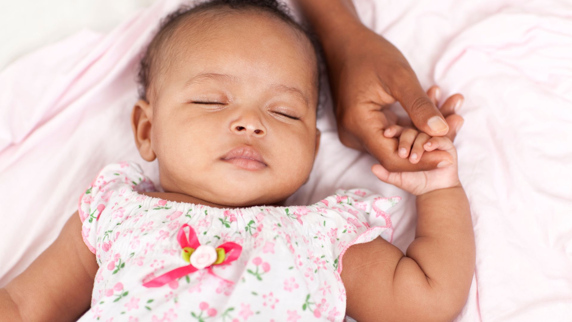 A baby sleeping on her back and her hand is wrapped around a mother’s finger. The baby is wearing a pink and flowery shirt.
