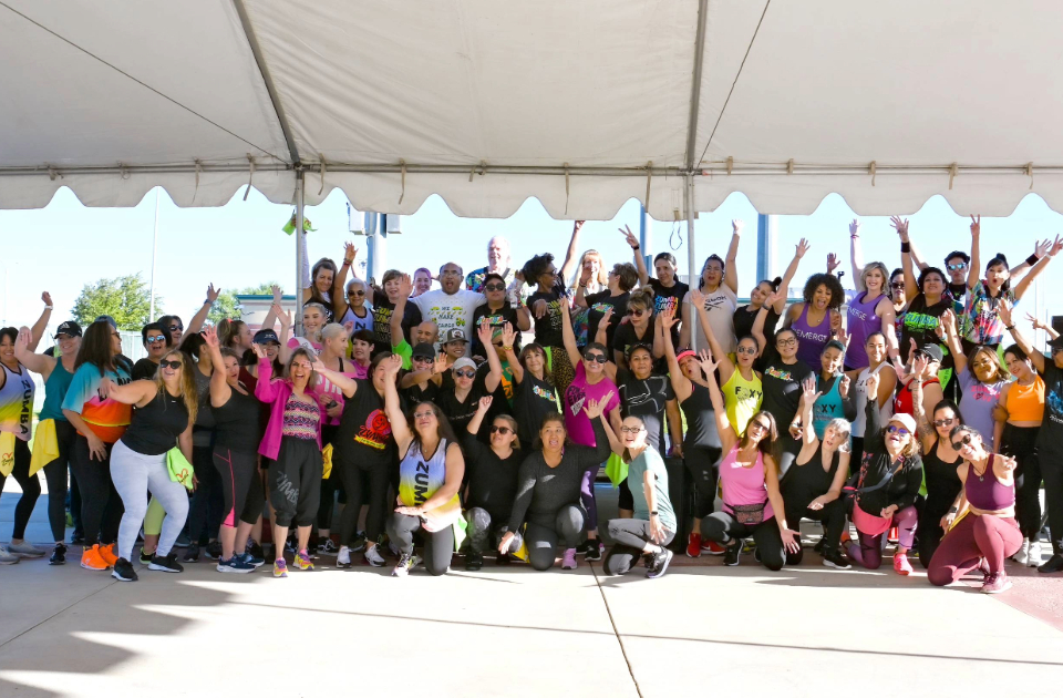 A group of over 50 people are smiling and raising their hands celebrating the end of a Zumbathon.